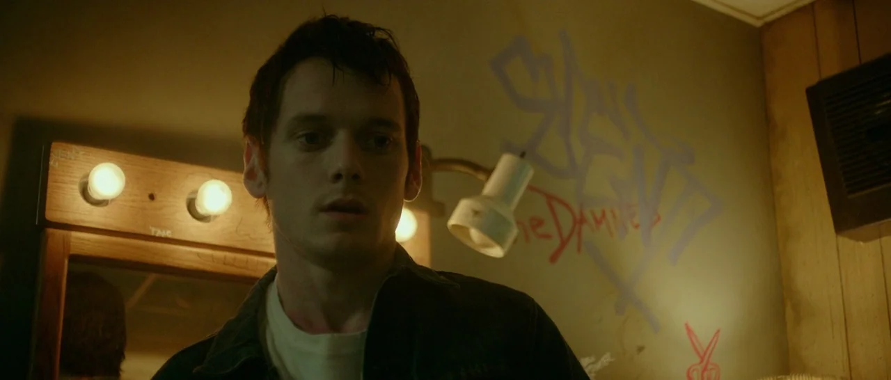 Green Room (2015) movie still: medium — A dimly lit room features a wooden vanity mirror and walls marked with spray-painted graffiti. A young man…