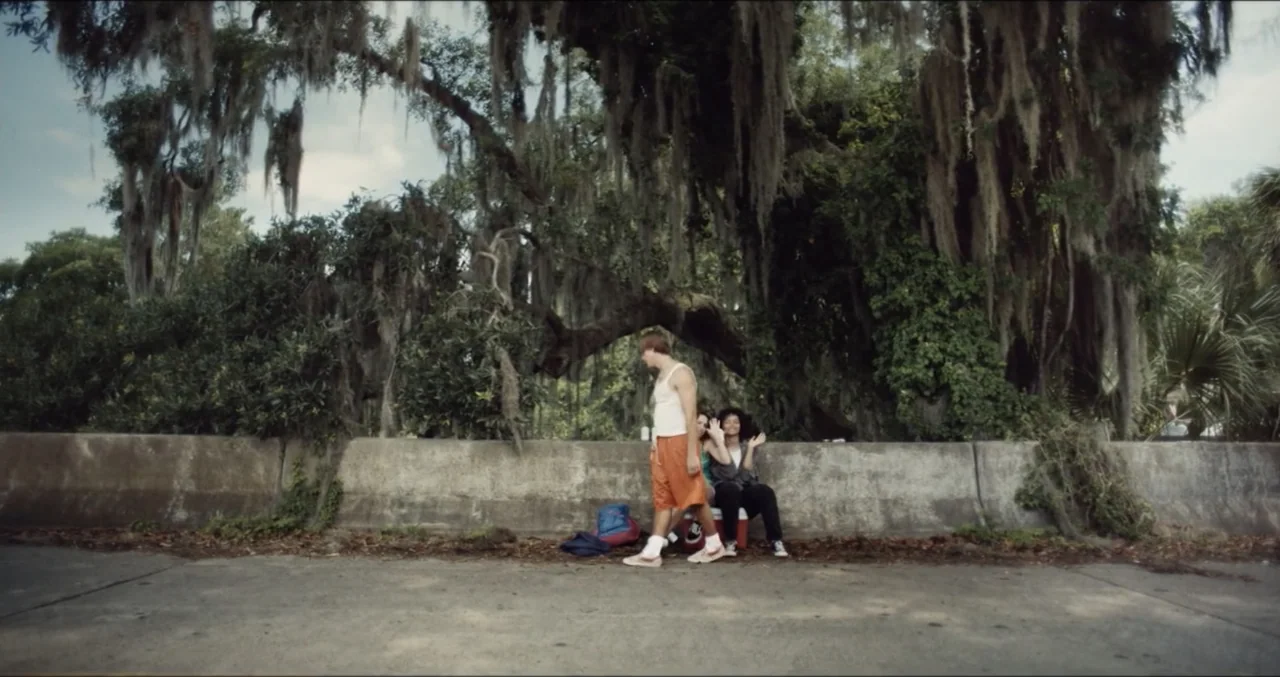 Eye for an Eye (2025) movie still: wide — An outdoor roadside area features a low concrete barrier beneath large Spanish moss trees. A young man…