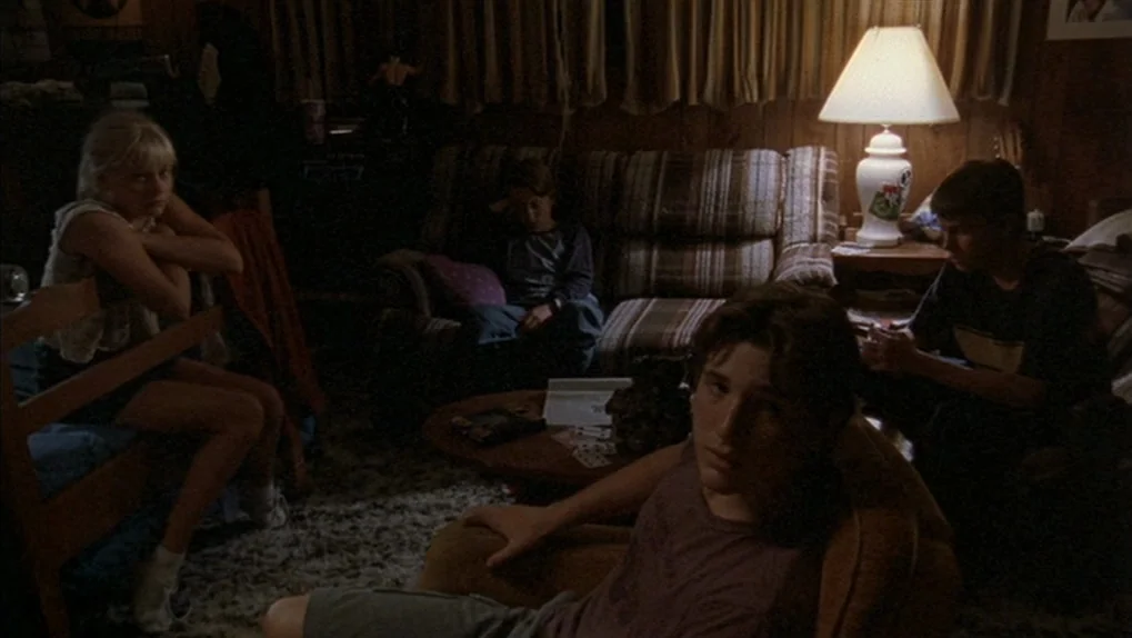 Mean Creek (2004) movie still: wide — A dimly lit 1990s living room features wood-paneled walls and a patterned couch. Four adolescents sit in…