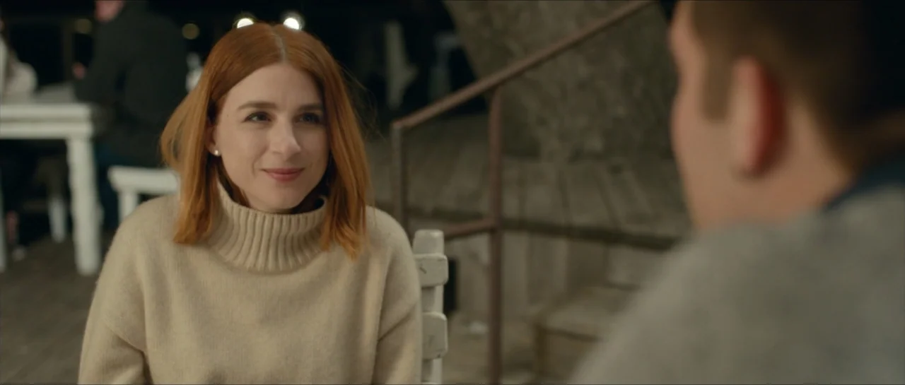 We Broke Up (2021) movie still: medium — An outdoor patio at night features a woman sitting at a table. She gazes toward a man whose back is turned…