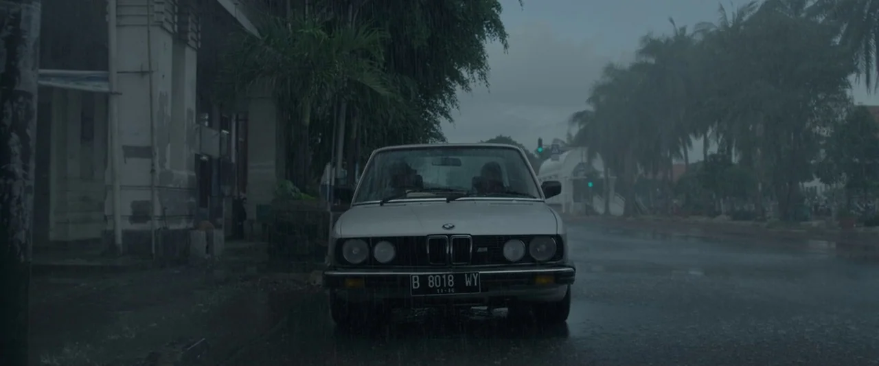 The Raid 2 (2014) movie still: medium — A silver vintage car is parked on a wet, rain-swept urban street. Palm trees and old building facades line…
