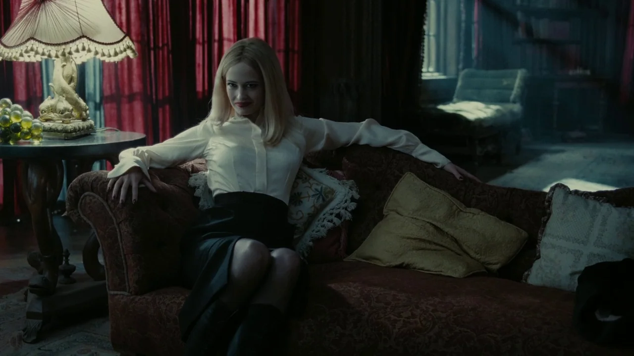 Dark Shadows (2012) movie still: medium — A dimly lit room features a velvet sofa and heavy dark red curtains. A blonde woman sits on the sofa,…