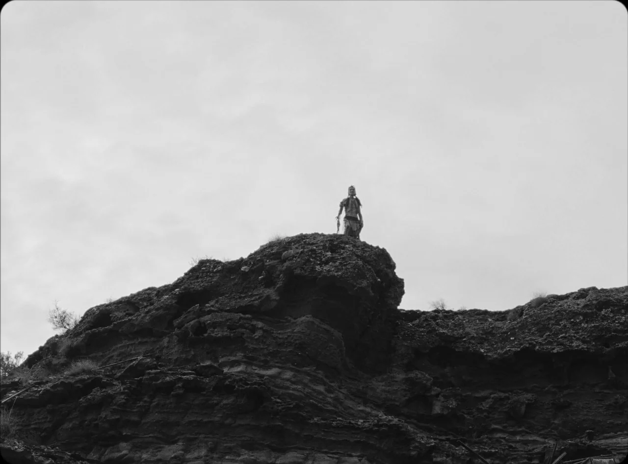 Eureka (2024) movie still: wide — A rugged dark rock formation dominates the frame against a bright, overcast sky. A single person stands at the…