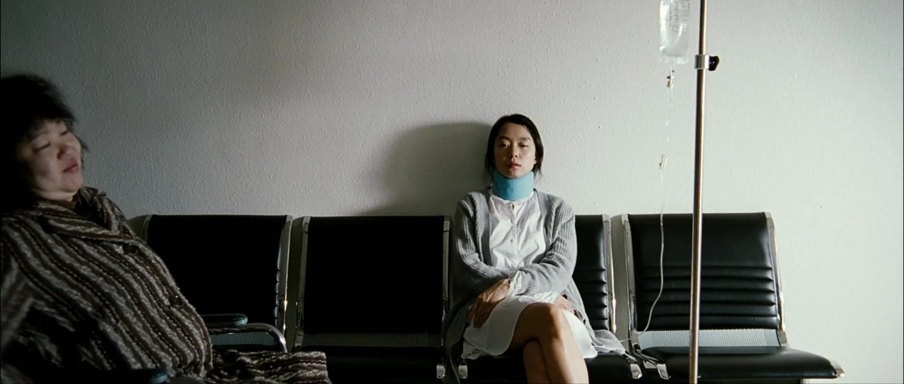 The Housemaid (2010) (2010) movie still: wide — A spartan hospital waiting area features black vinyl chairs against a blank white wall. A young…