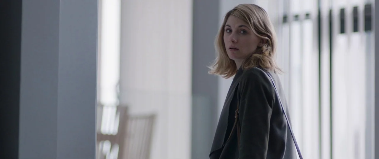 Journeyman (2017) movie still: medium — A clean, modern corridor features large floor-to-ceiling windows and cool-toned gray wall surfaces. A woman…