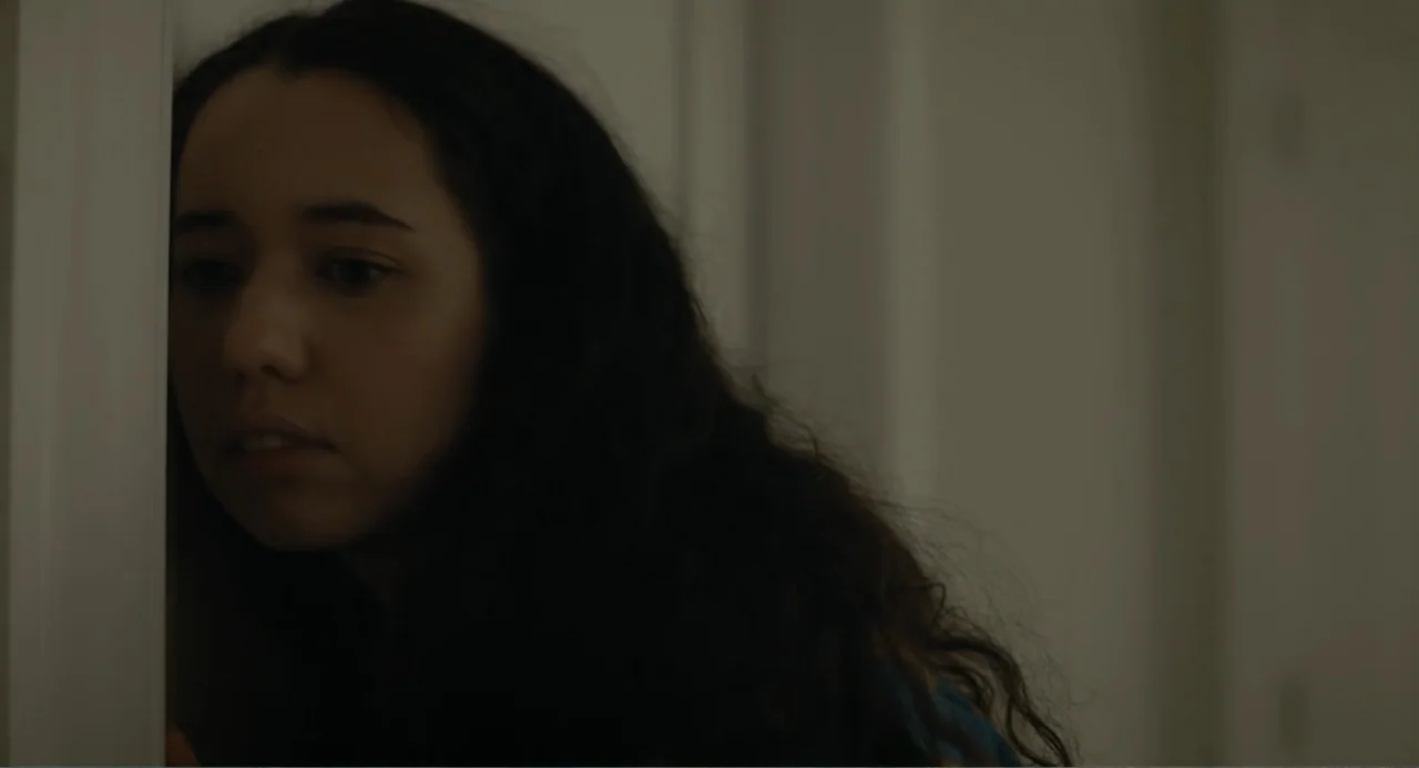 This Closeness (2024) movie still: closeup — A dark indoor doorway is partially framed by a white vertical wall element. A young woman with long…