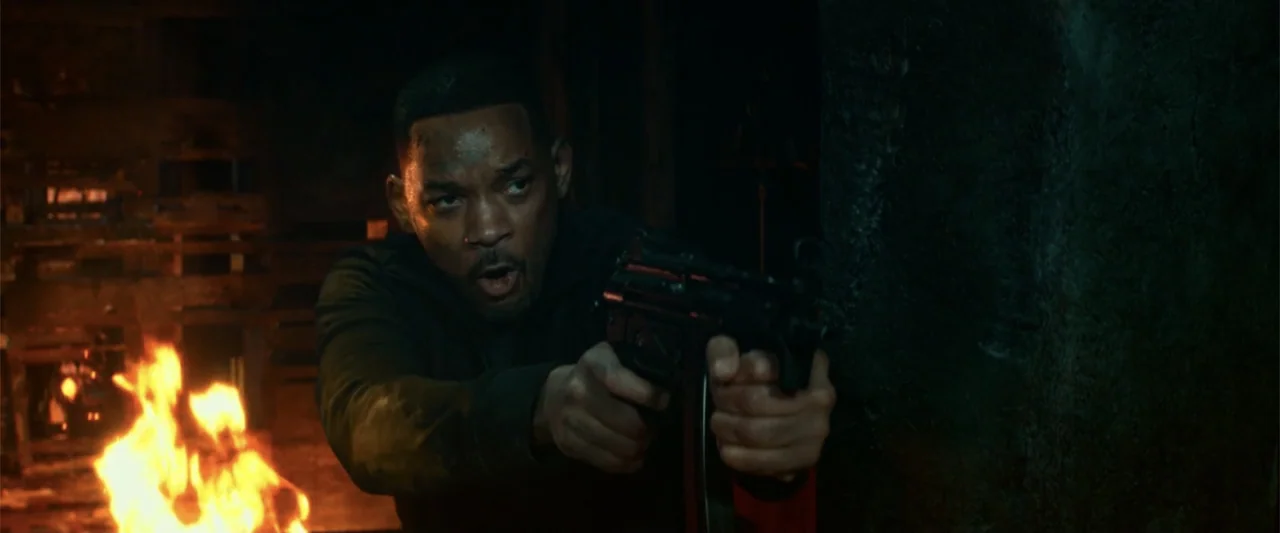 Bad Boys for Life (2020) movie still: medium — A dark industrial interior with flames burning in the background. A man holds a firearm while looking…
