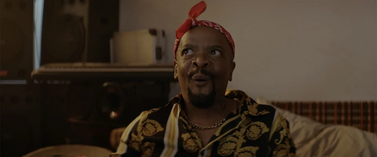 A Scam Called Love (2025) movie still: medium — A dimly lit room contains audio equipment and speakers in the background. A man wears a red bandana…