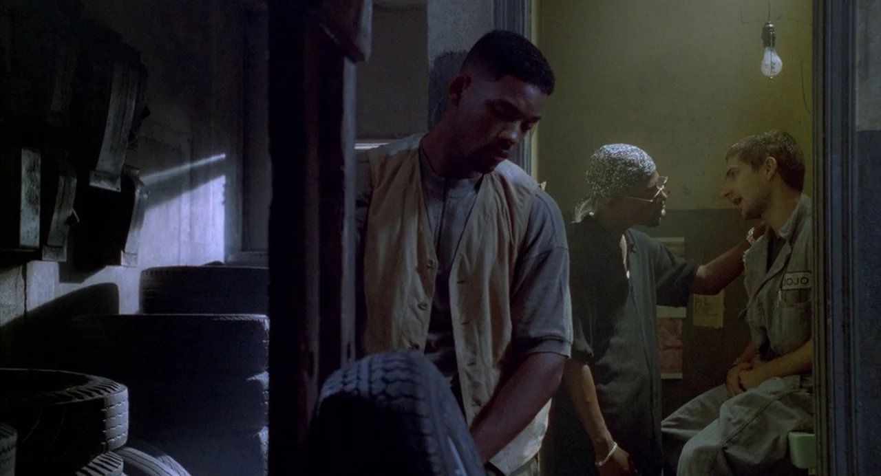 Bad Boys (1995) movie still: medium — A dark, cluttered garage features stacks of tires along a dim brick wall. Two men have a conversation while a…