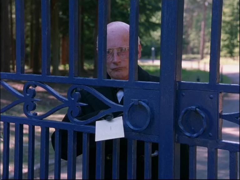 Eyes Wide Shut (1999) movie still: medium — A blue metal gate stands in the foreground, obscuring a wooded outdoor area. An elderly man looks…
