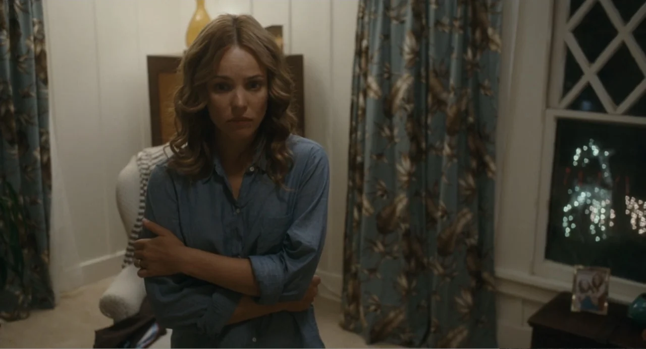 Aloha (2015) movie still: medium — A residential bedroom interior features wood paneled walls, patterned curtains, and a window view. A woman stands…