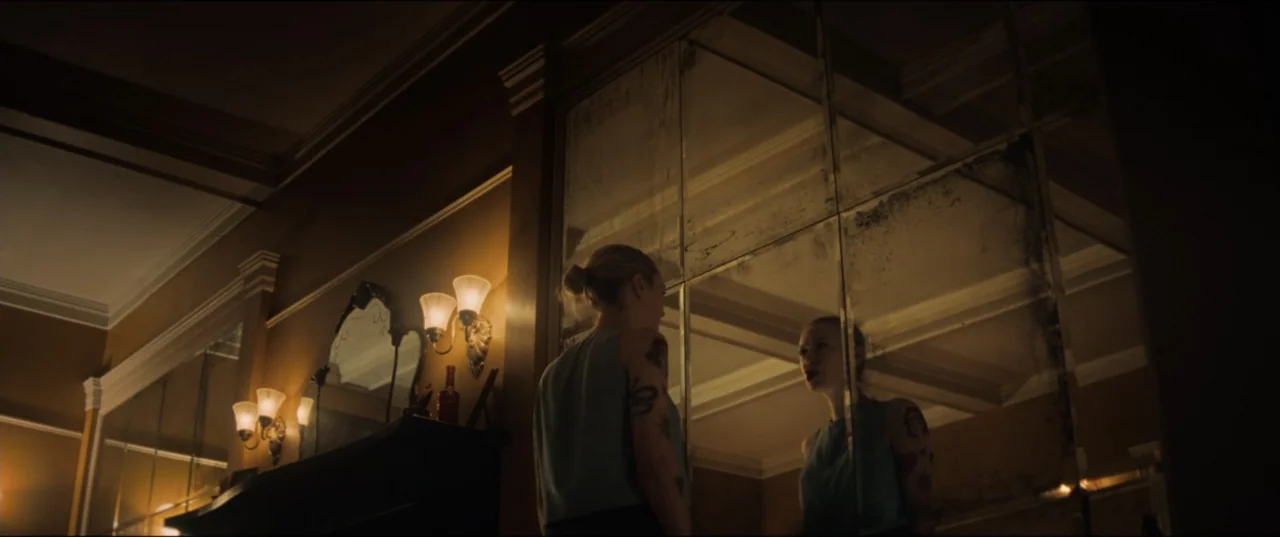 Vicious (2025) movie still: medium — A dimly lit room features a wall covered in large, aged, multi-pane mirrors. A young woman with tattoos on her…