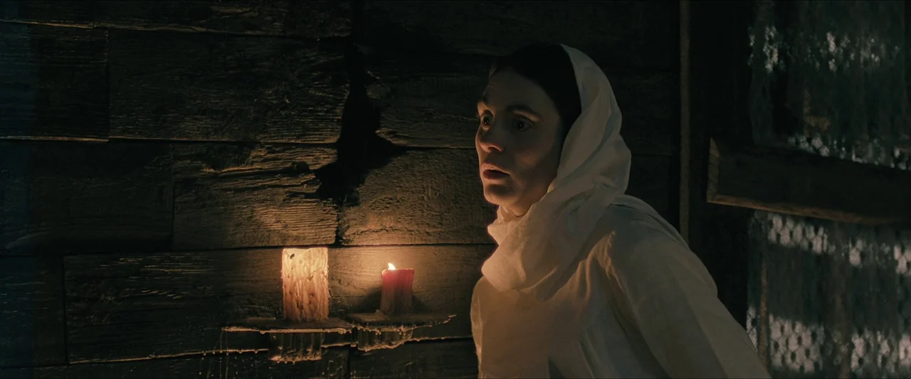 Azrael (2024) movie still: medium — A dark, cramped room is constructed from rough-hewn horizontal timber logs. A young woman in a white headscarf…
