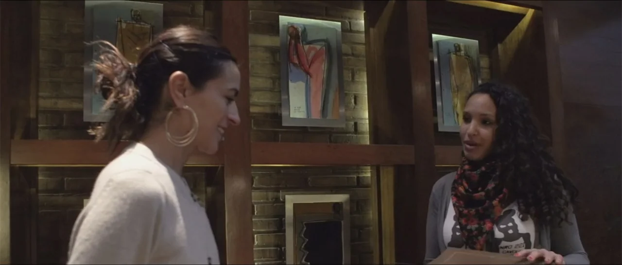 10 Years with Bebe (2016) movie still: medium — An interior space features wood shelving units holding framed abstract paintings against brick…