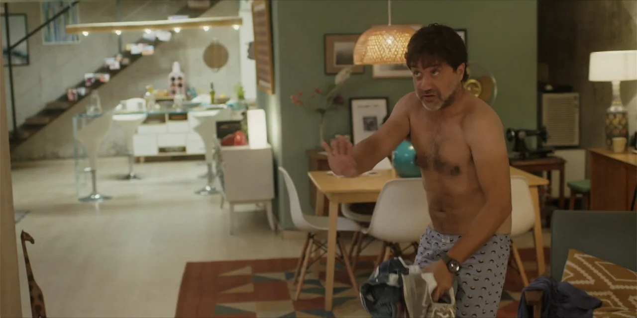 Instant Love (2020) movie still: medium — A modern apartment interior features a central dining area and a staircase. A shirtless man stands in the…