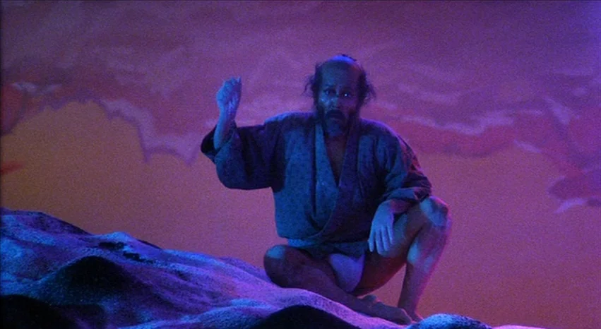 Kagemusha (1980) movie still: medium — A theatrical stage set features a stylized, painted backdrop with dramatic purple lighting. An elderly man in…