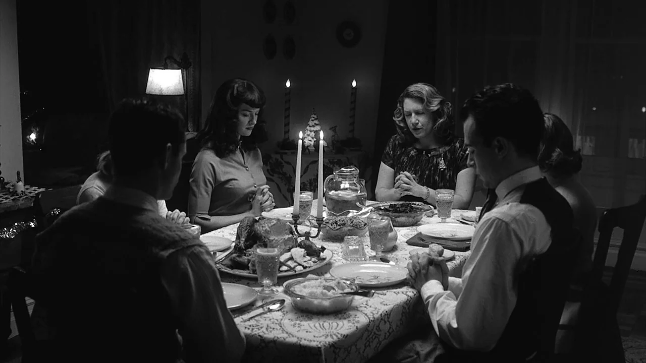 The Notorious Bettie Page (2005) movie still: medium — A 1950s dining room features a table set with a large meal for dinner. Four people sit around…
