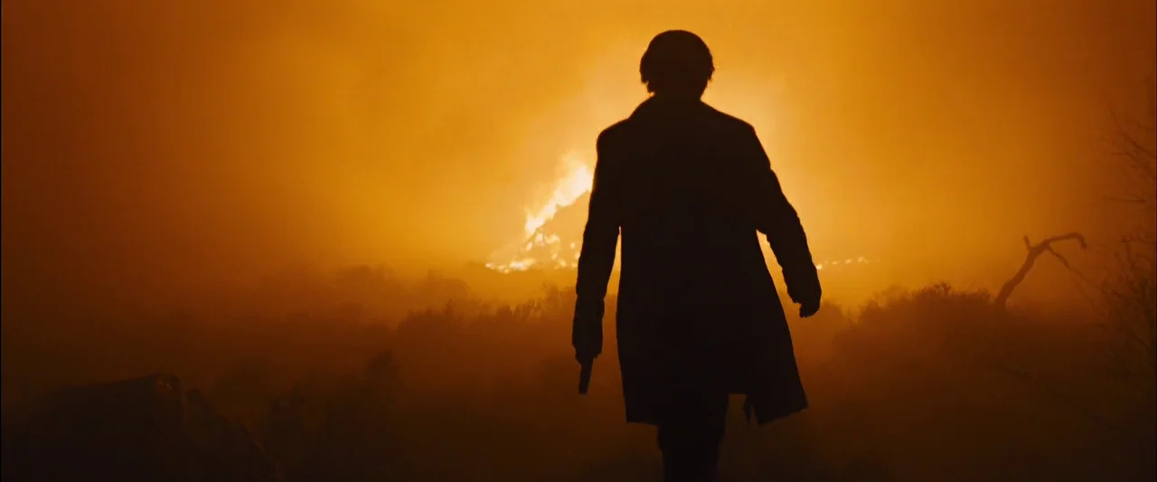 Skyfall (2012) movie still: fullbody — A field is obscured by thick orange smoke and glowing embers from a wildfire. A man in a long coat walks away…