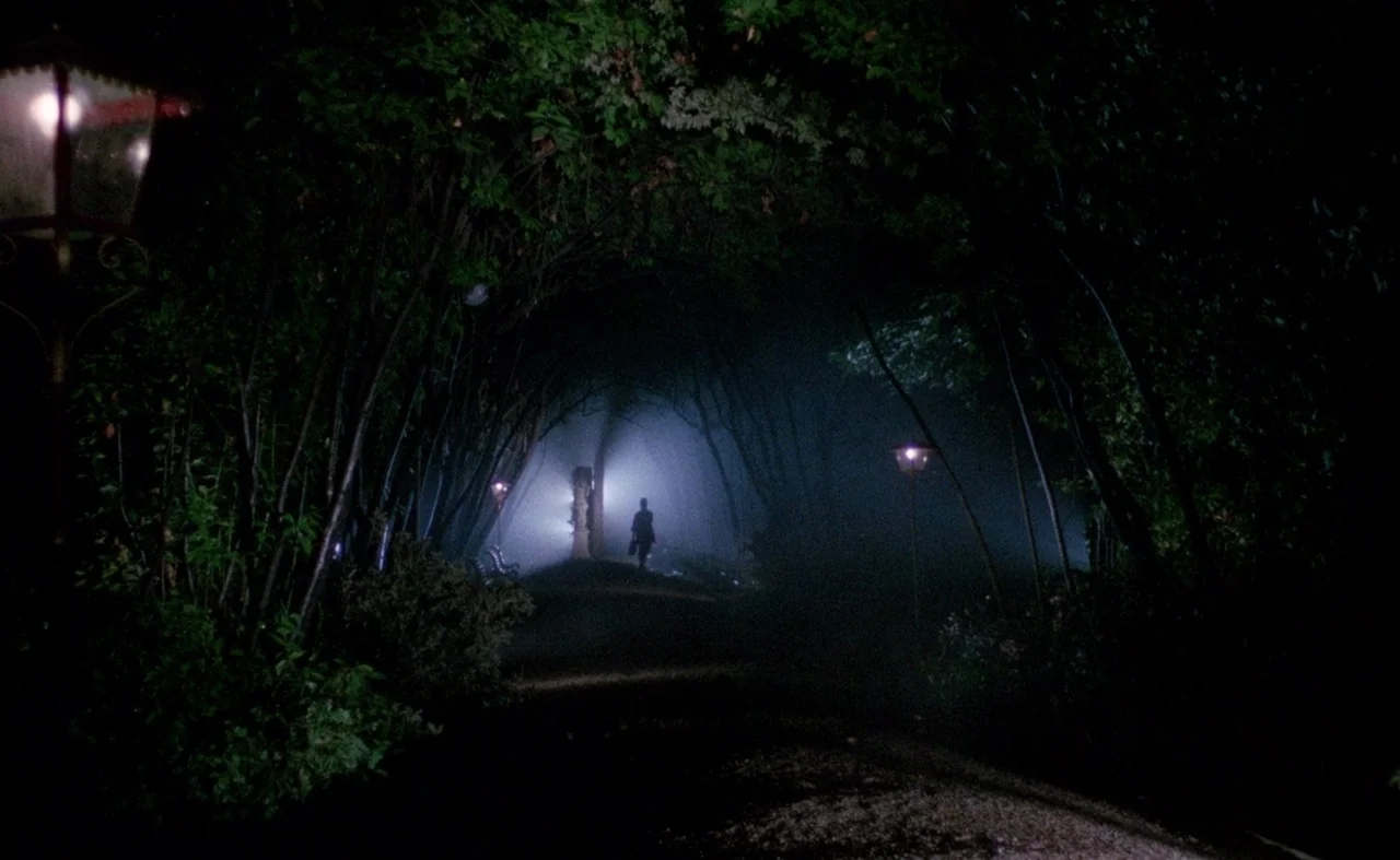 Blood And Black Lace (1964) movie still: wide — A dark, tree-lined park path is shrouded in a thick, low-lying mist at night. A lone silhouette…