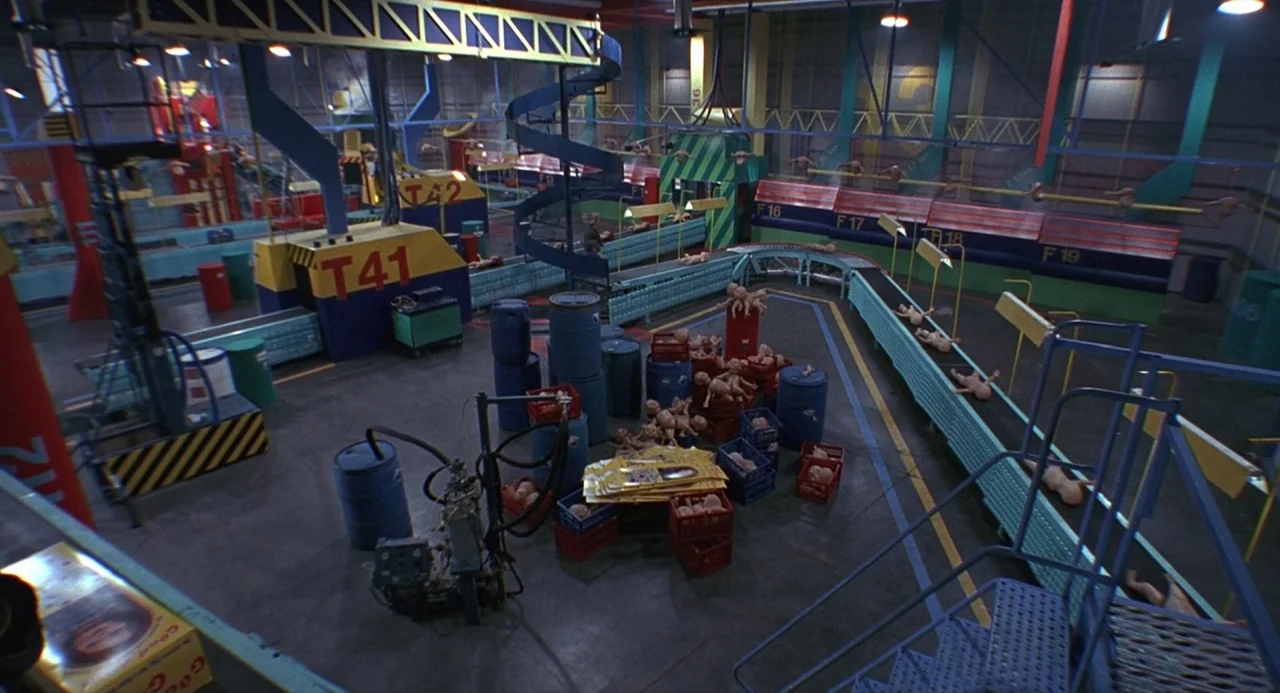 Child’s Play 2 (1990) movie still: wide — A sprawling toy factory floor features yellow and blue machinery and automated conveyor belts. Many…