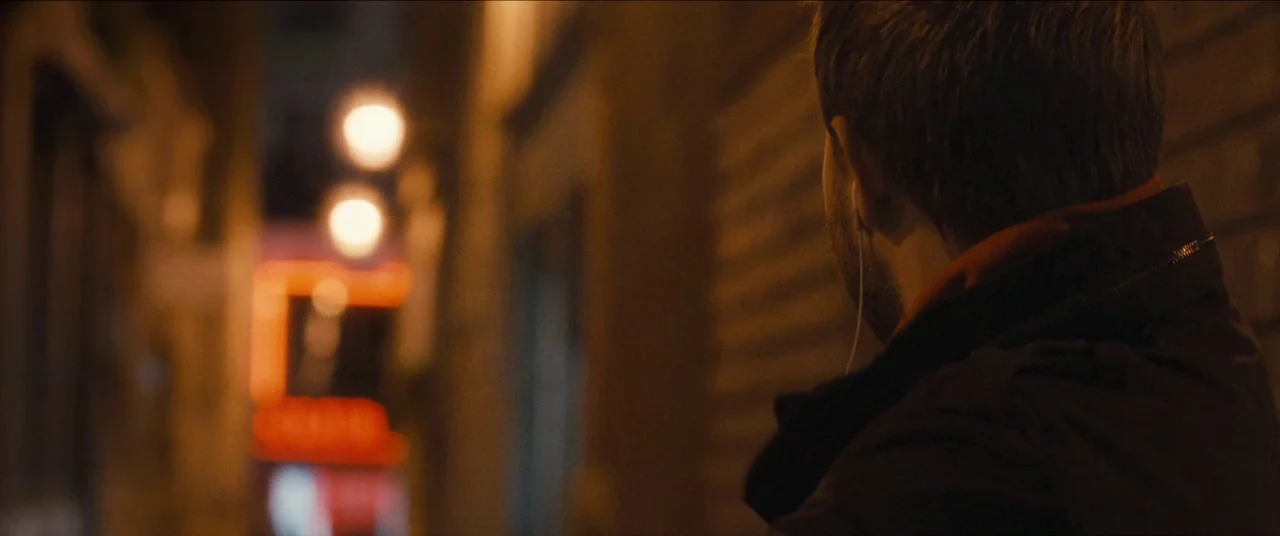Simon Killer movie still: medium — A dark, narrow urban walkway at night with blurred orange neon lights in distance. A young man seen from behind…