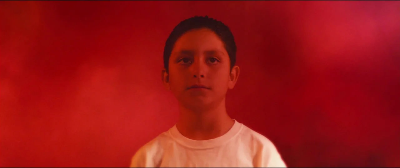 El Chicano (2018) movie still: closeup — A young boy stands in an abstract space filled with deep red light. The boy looks directly toward the…