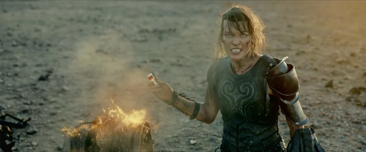 Monster Hunter (2020) movie still: medium — An open rocky landscape features a small fire smoldering on the dry ground. A woman in dark leather…