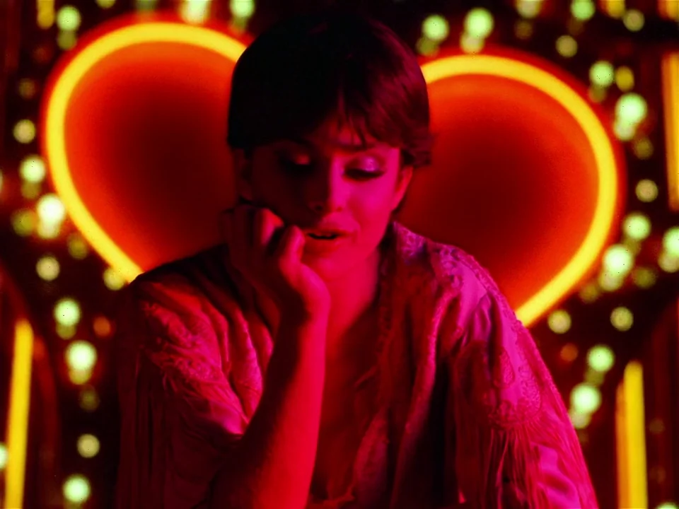 One From The Heart (1982) movie still: medium — A dark, dimly lit environment features a prominent neon sign shaped like a heart. The woman sits…