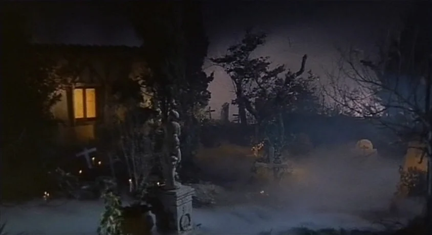 Kill, Baby Kill (1966) movie still: establishing — A dark, mist-filled cemetery at night features gravestones, sparse trees, and a lit window. Thick…