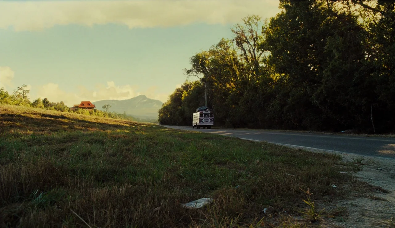 Our Brand is Crisis (2015) movie still: wide — A rural asphalt road cuts through a grassy meadow bordered by a dense tree line. A small bus drives…