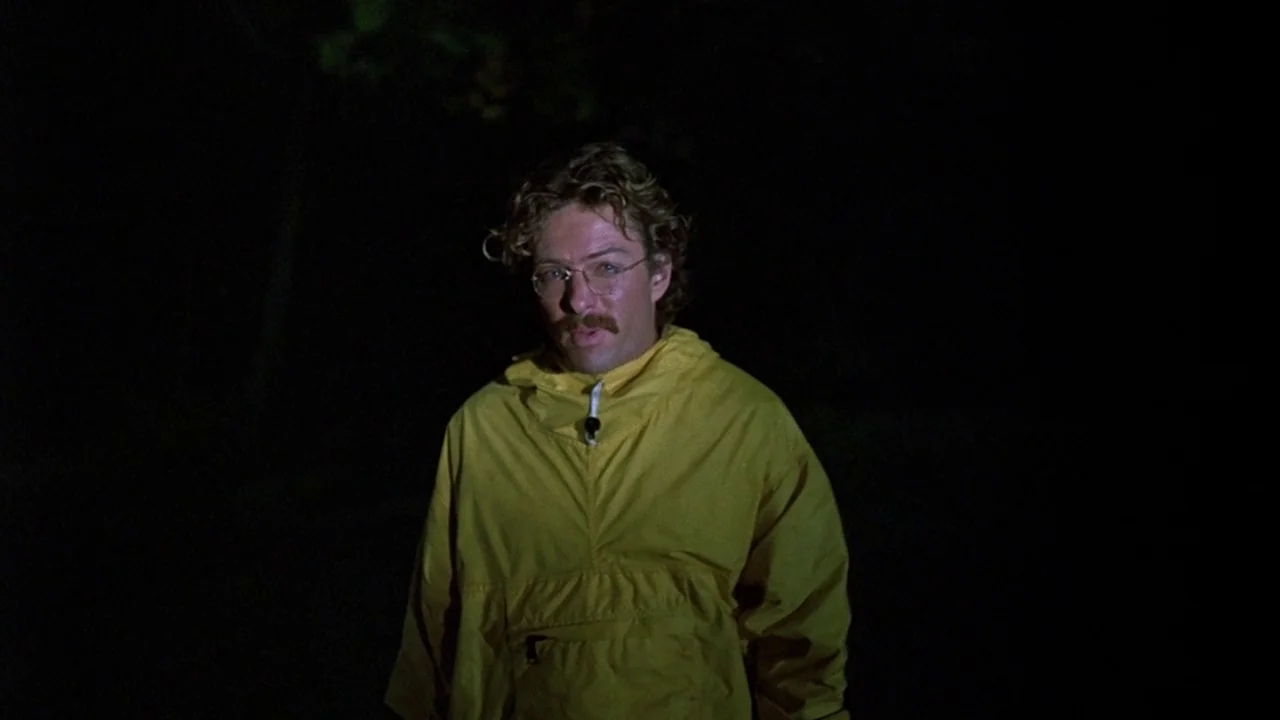 Friday the 13th (1980) movie still: medium — The man stands in an outdoor environment completely shrouded in darkness. A bright yellow jacket stands…