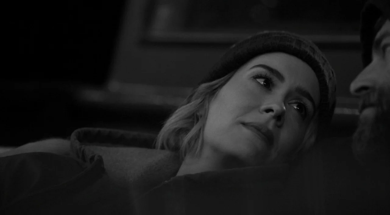 Blue Jay (2016) movie still: closeup — A car interior at night with low lighting levels obscuring most of the cabin. A woman in a beanie rests her…