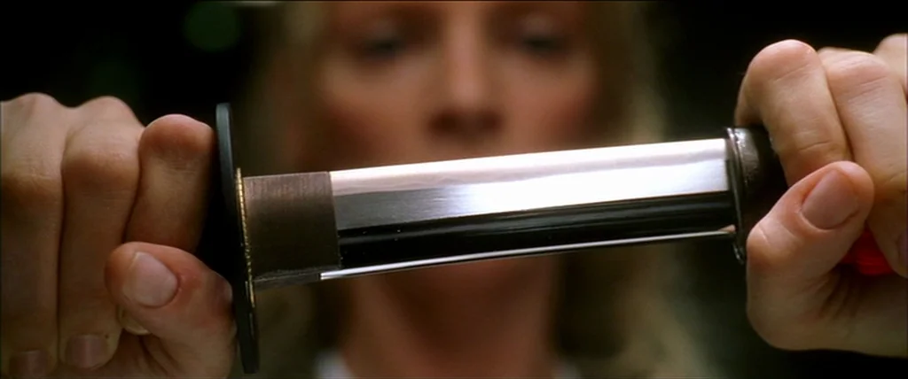 Kill Bill Vol. 1 (2004) movie still: closeup — A close-up of a sharp steel katana blade held firmly by two hands. The background is a soft-focus…