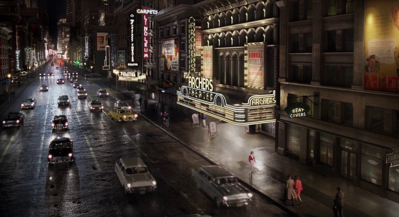 Populaire (2012) movie still: establishing — A rain-slicked city street features an ornate theater marquee with prominent neon signage at night.…