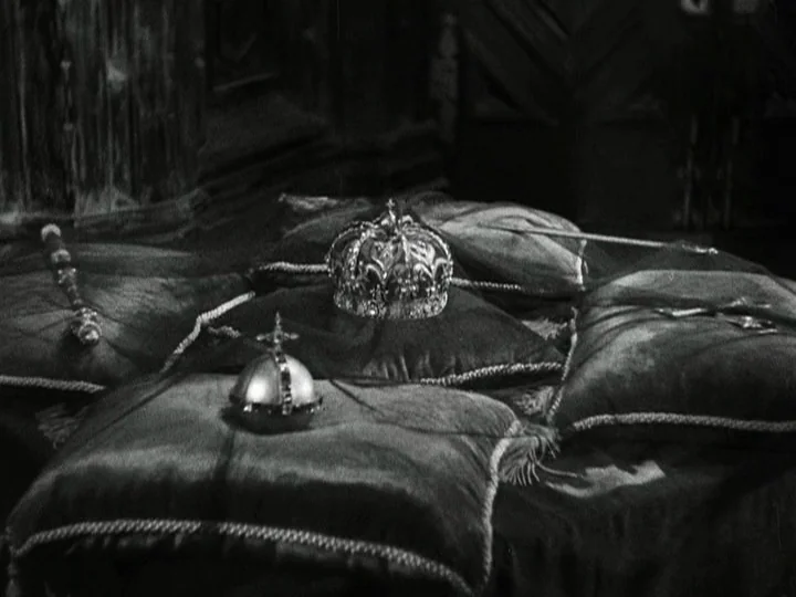Queen Christina (1933) movie still: closeup — A collection of royal regalia rests upon velvet cushions in a dimly lit chamber. A crown, orb, and…