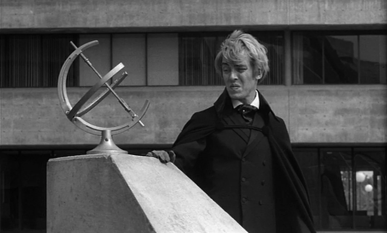 Stereo (1969) movie still: medium — A man stands before a concrete structure in front of a brutalist building facade. He rests his gloved hand on a…