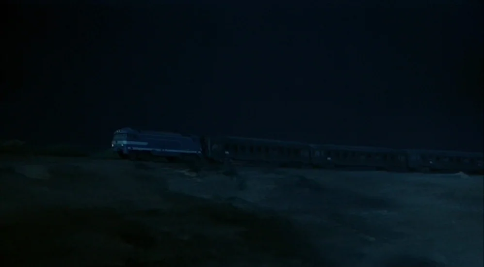 Un Flic (1972) movie still: wide — A dark, empty landscape at night features a blue train moving along tracks. The train consists of a locomotive…