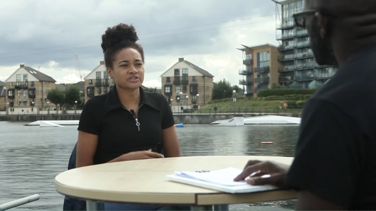 A Film Called Blacks Can't Swim (My Journey My Story) (2020) movie still: medium — A woman sits at a round table placed on a dock near water. She…