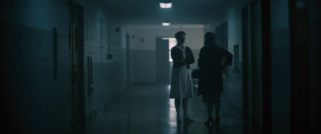 Boston Strangler (2023) movie still: wide — A dimly lit, long institutional hallway with tiled walls and overhead fluorescent lights. Two women in…