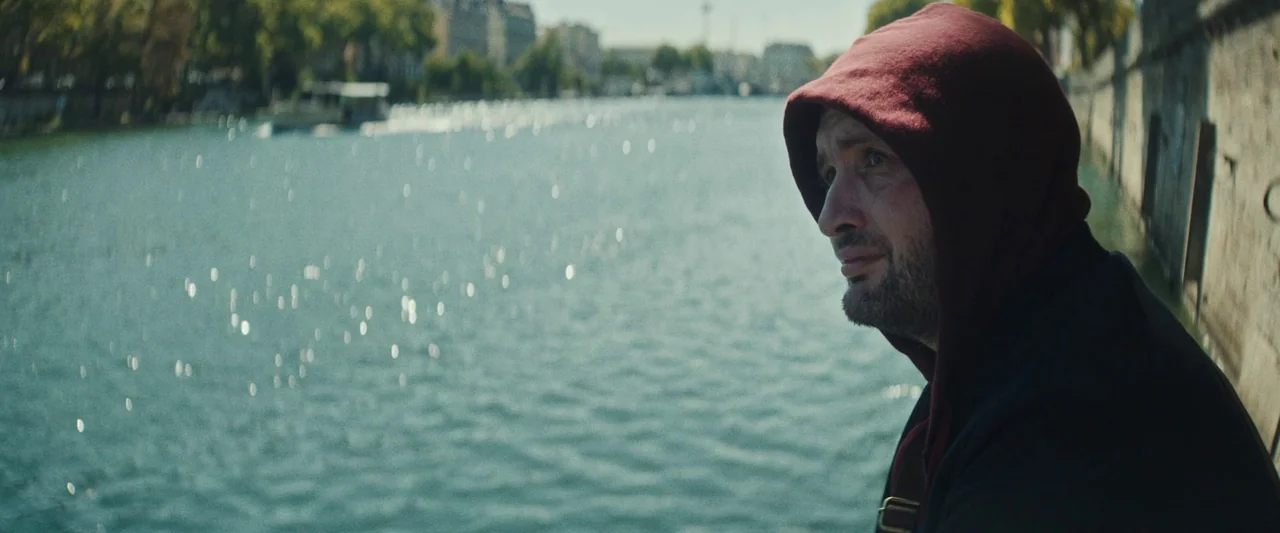 Vincent Must Die (2023) movie still: medium — A man stands by a stone wall overlooking a wide river in a city. He watches the water while wearing a…