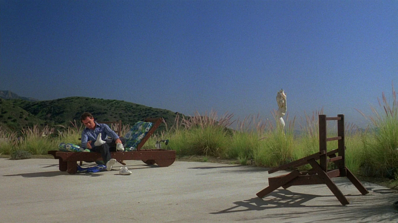 Paris, Texas (1984) movie still: wide — A concrete patio sits at the edge of a dry grassy hill under blue sky. A man in a striped shirt sits on a…