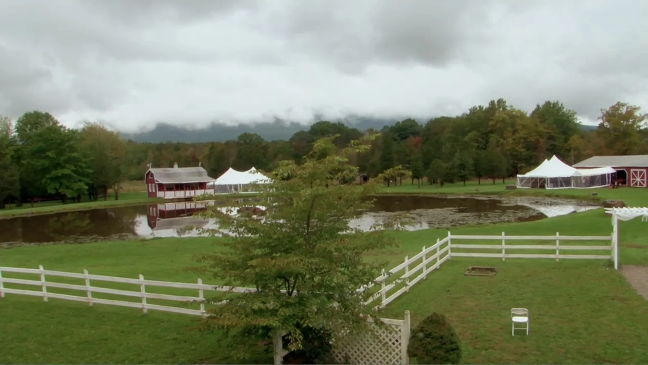 Little White Lie (2008) movie still: establishing — A rural grassy clearing features a small pond with white event tents nearby. A red wooden barn…
