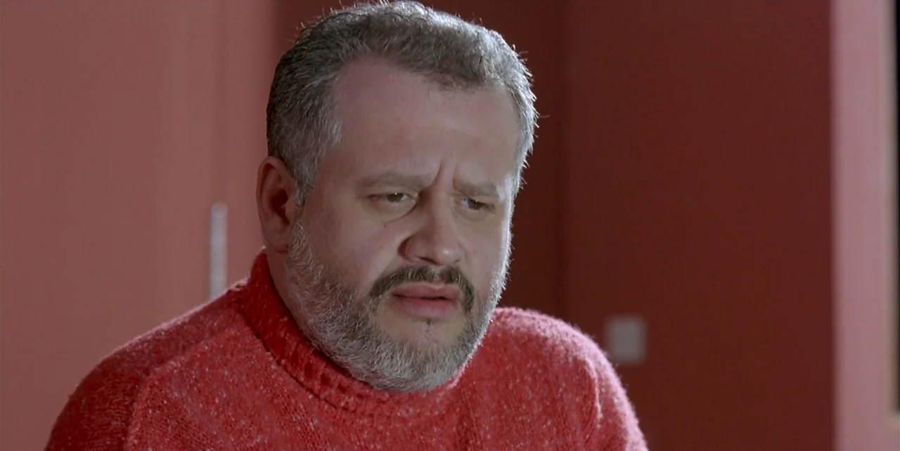 The Flower of My Secret (1995) movie still: closeup — The interior space features plain walls in a warm red tone. A middle-aged man with a beard…