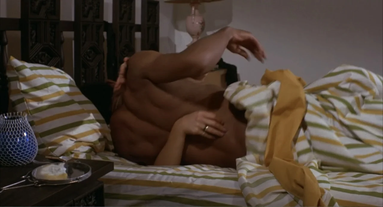 Foxy Brown (1974) movie still: medium — A residential bedroom features a wooden bed frame and striped white, green, and yellow linens. Two people…