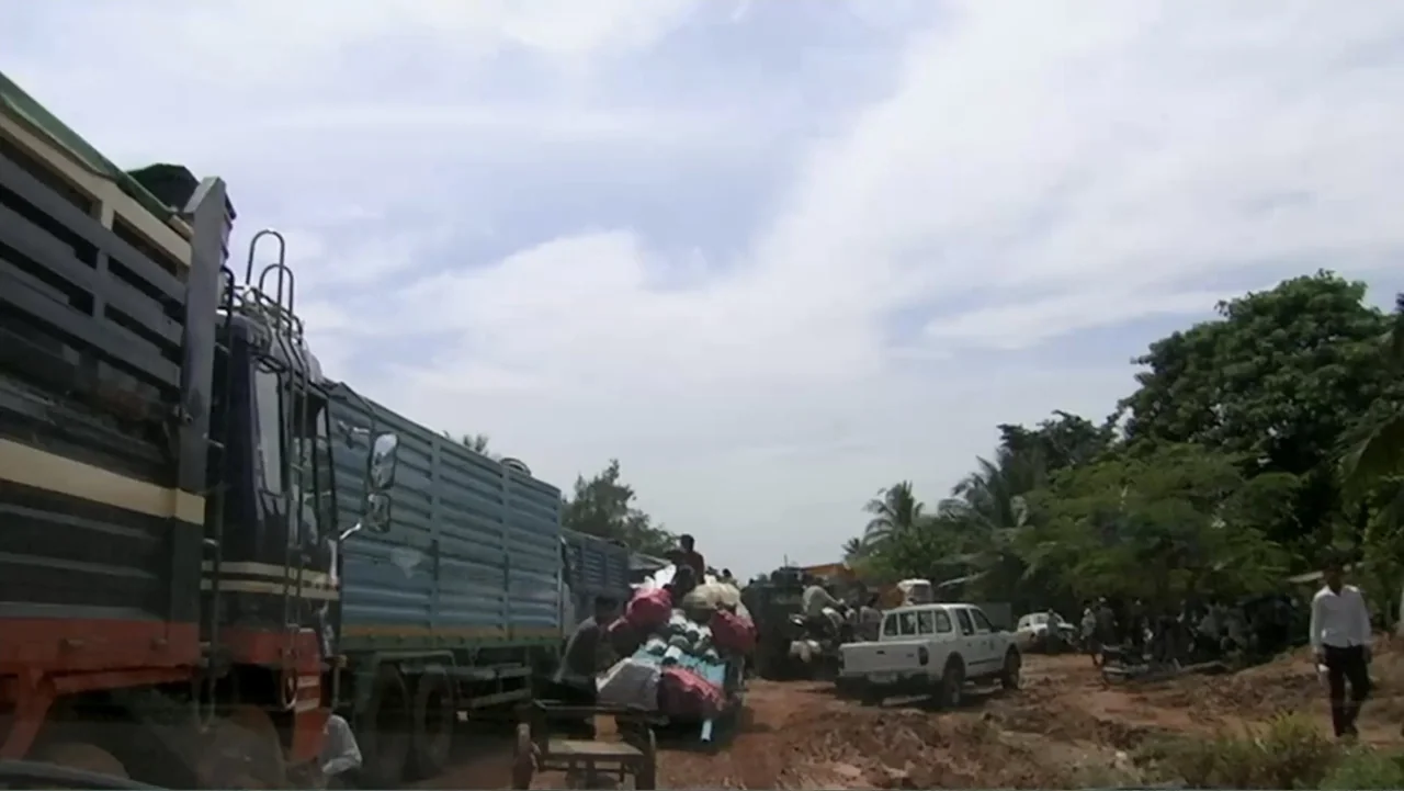 Sister (2014) movie still: wide — A muddy, unpaved road flanked by lush green vegetation and parked heavy vehicles. People are clustered near trucks…