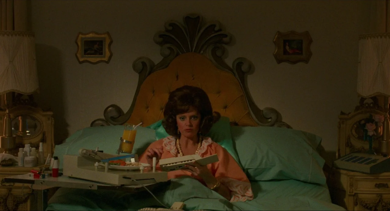 True Stories (1986) movie still: medium — A woman sits propped up in a plush hotel bed with turquoise linens. She works with a portable typing…