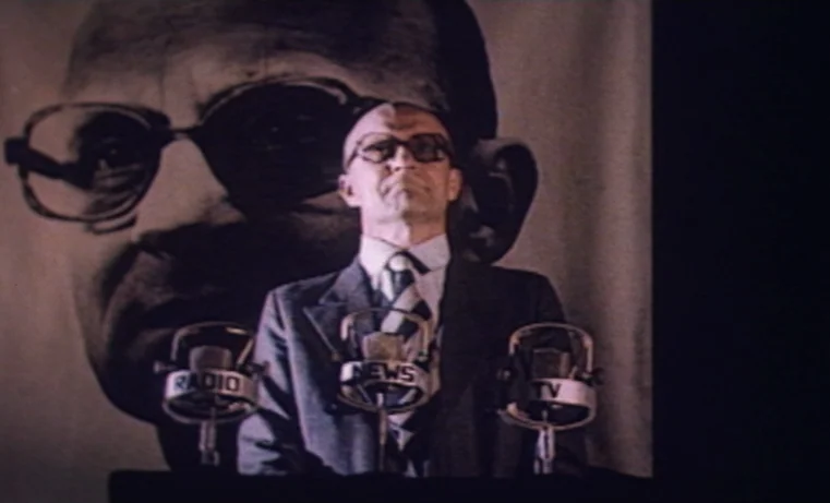 Golem (1980) movie still: medium — A man stands at a podium in front of a giant poster of a person. Three vintage radio and news microphones are…