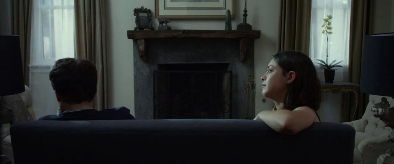 Night Owls (2015) movie still: medium — A living room interior featuring a central stone fireplace topped with a wooden mantel. A man and woman sit…