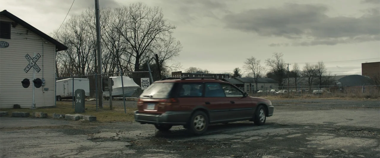 Skin (2009) movie still: wide — A rural railroad crossing with a gravel lot and a chain-link fence. An old red station wagon drives across the…