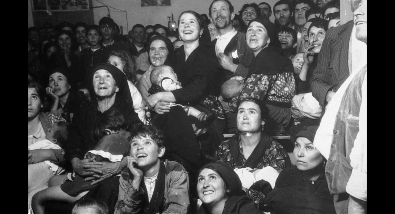 Los días azules (2025) movie still: medium — A crowded interior room is filled with seated men, women, and small infants. The group looks toward an…